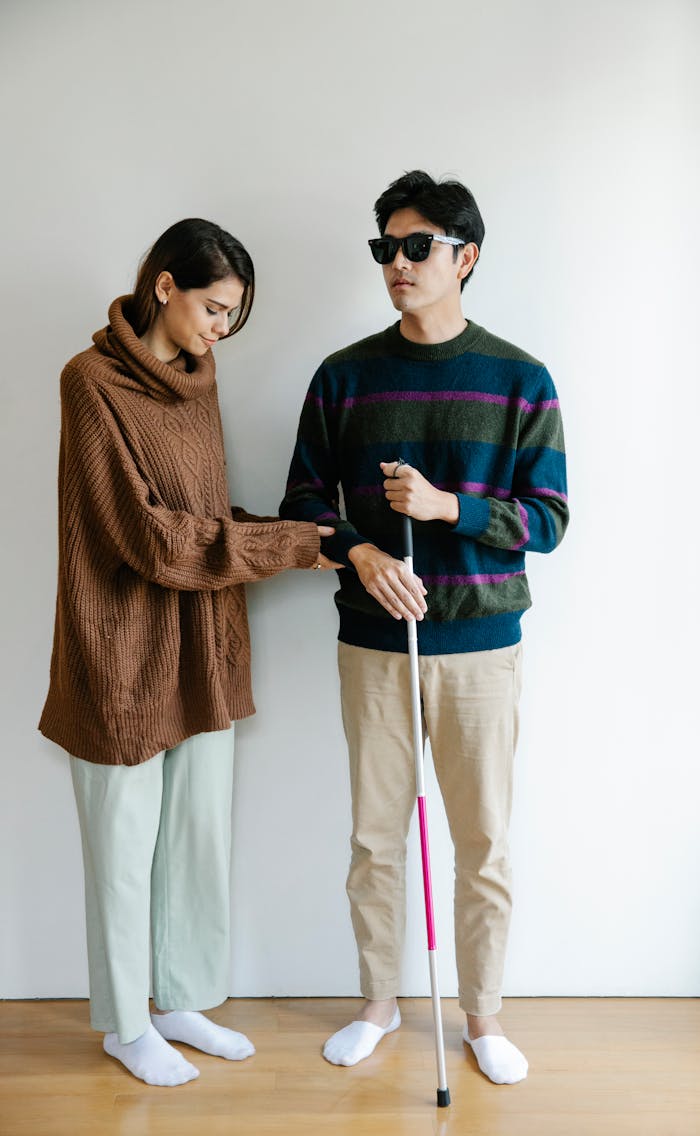 A woman helping a visually impaired man with a walking stick indoors.