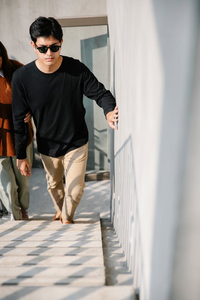 A man with visual impairment uses touch for support while ascending stairs, guided by a companion.